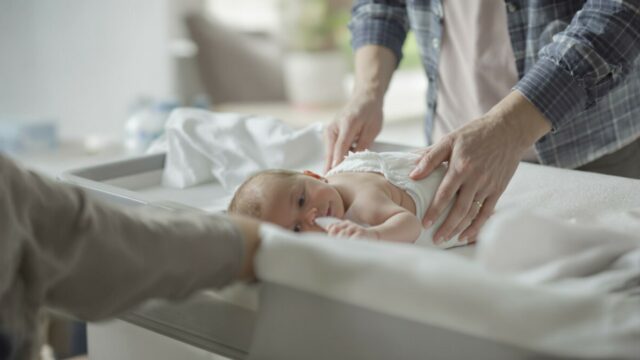 Un bébé sur une table à langer se fait changer sa couche par deux adultes.